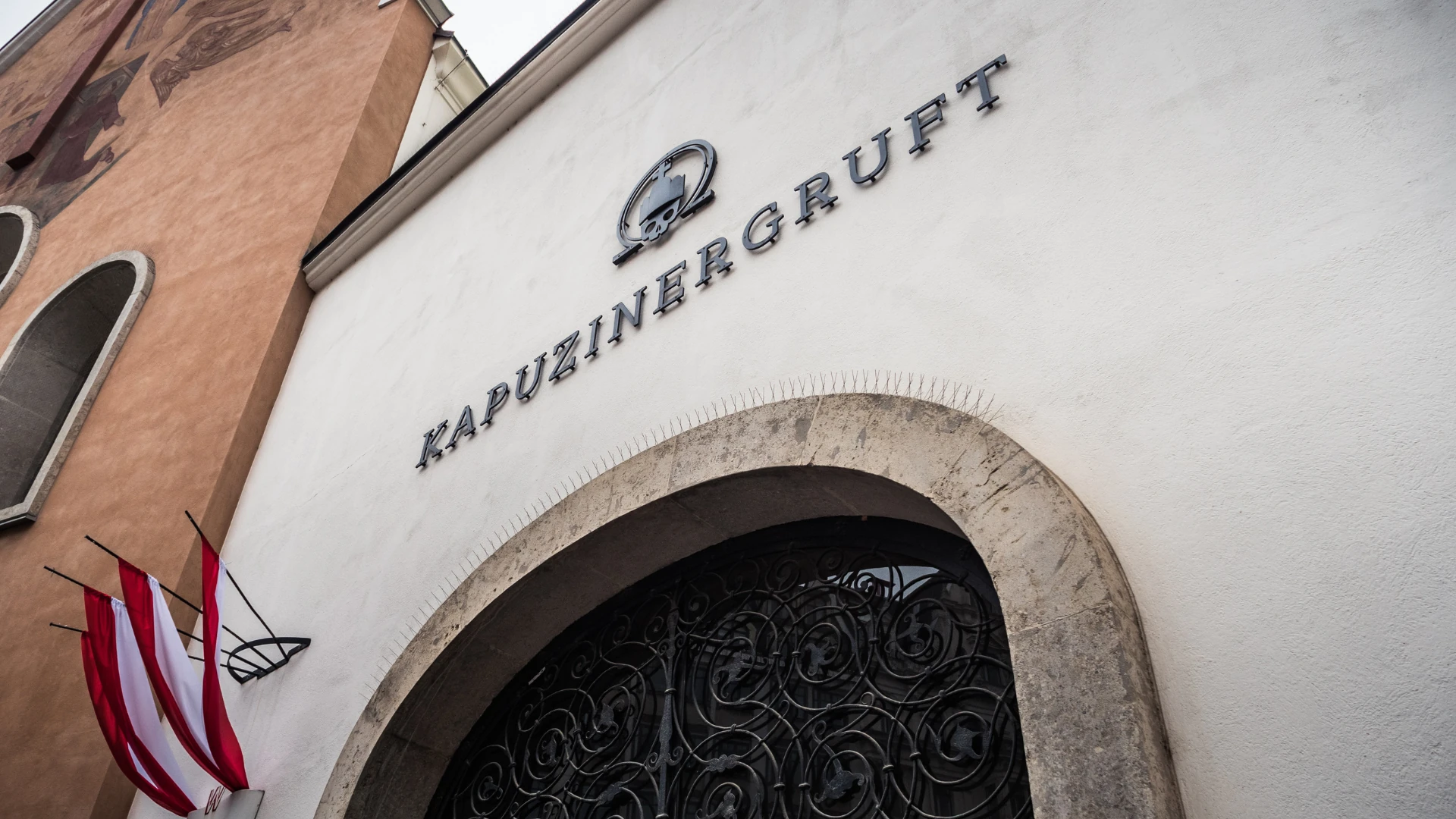 Dieses Foto zeigt den Eingang zur Kapuzinergruft in Wien, auch bekannt als Kaisergruft. Die schlichte weiße Fassade mit dem markanten Schriftzug und den österreichischen Fahnen verweist auf die letzte Ruhestätte der Habsburger-Dynastie. Das kunstvolle schmiedeeiserne Tor unterstreicht die historische Bedeutung dieses Ortes.