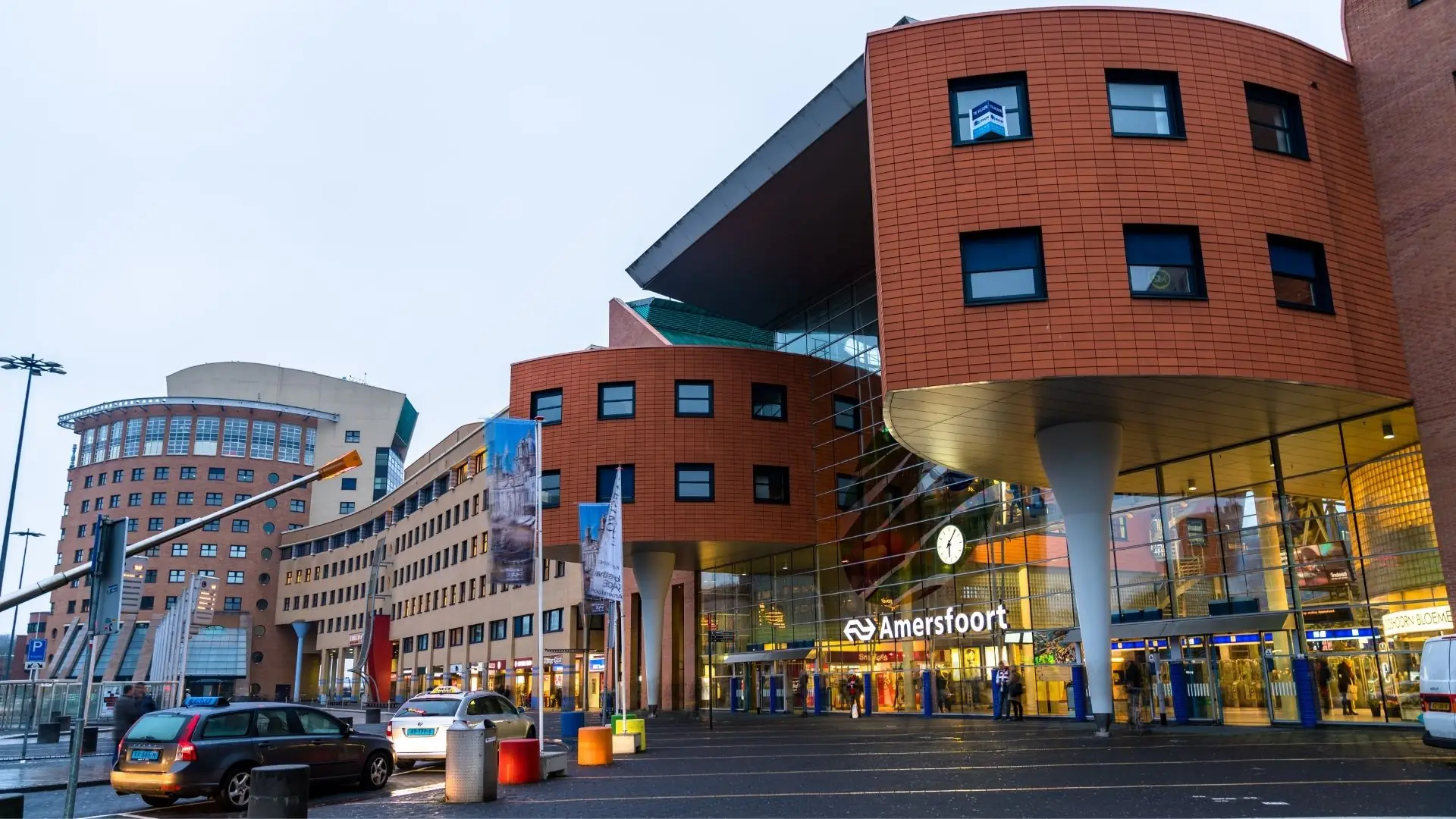 Außenansicht des Bahnhofs Amersfoort Centraal mit rotem Backsteingebäude, mehreren Fenstern und beleuchtetem Eingangsbereich bei Tageslicht.