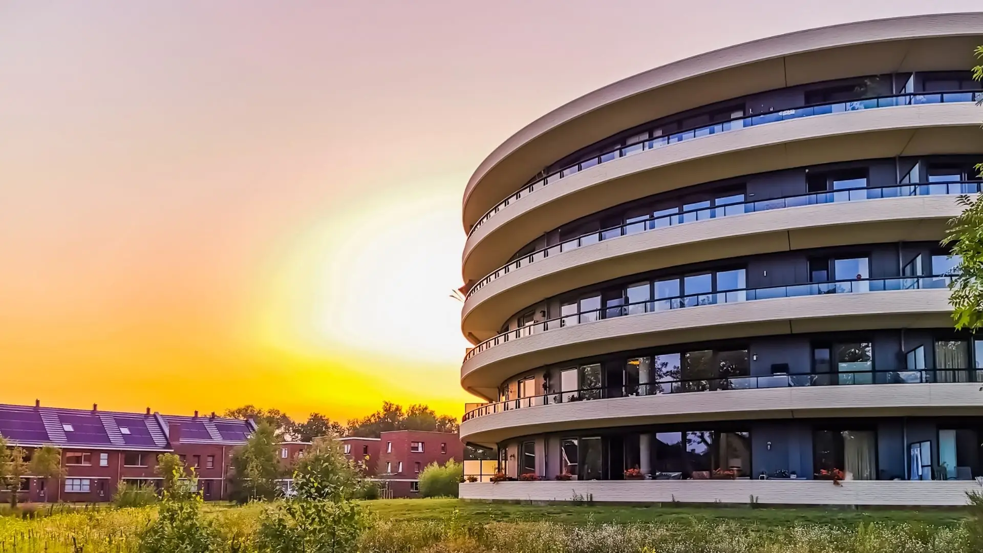 Rundes, mehrstöckiges Wohngebäude mit Balkonen neben Reihenhäusern und Bäumen bei Sonnenuntergang.