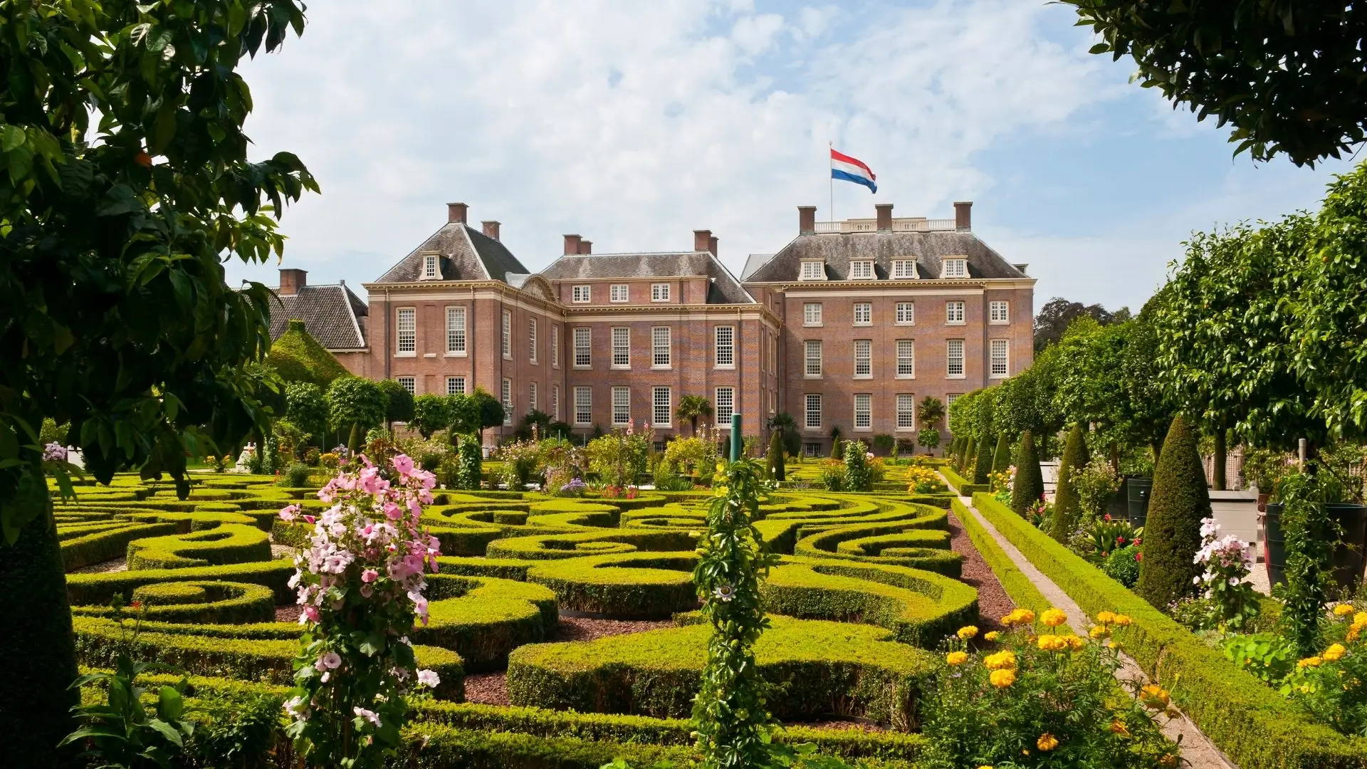 Das Paleis Het Loo mit einer niederländischen Flagge auf dem Dach, davor ein symmetrischer Garten mit grünen Hecken und bunten Blumen.