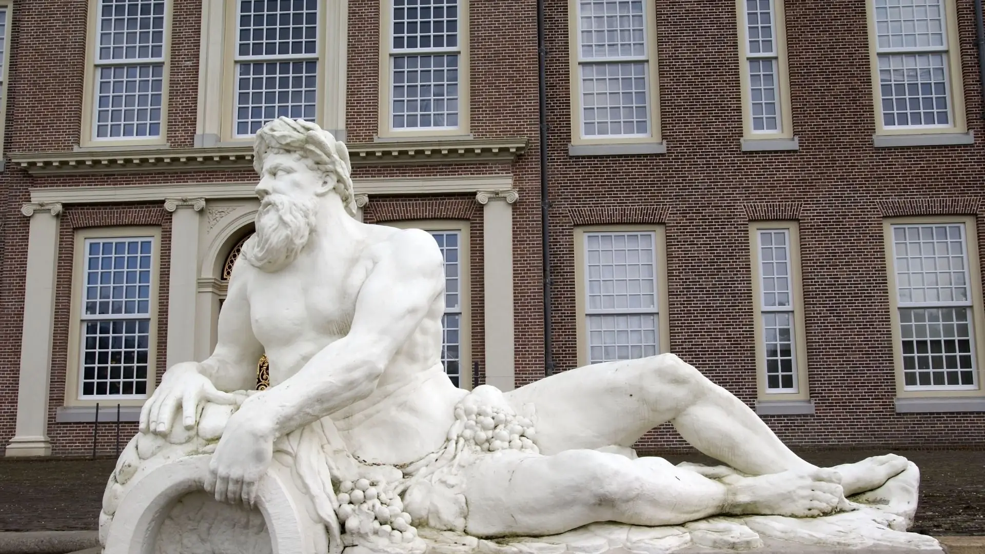 Weiße Marmorstatue eines liegenden, muskulösen Mannes mit langem Haar, umgeben von Weintrauben, vor einem Gebäude mit großen Fenstern.