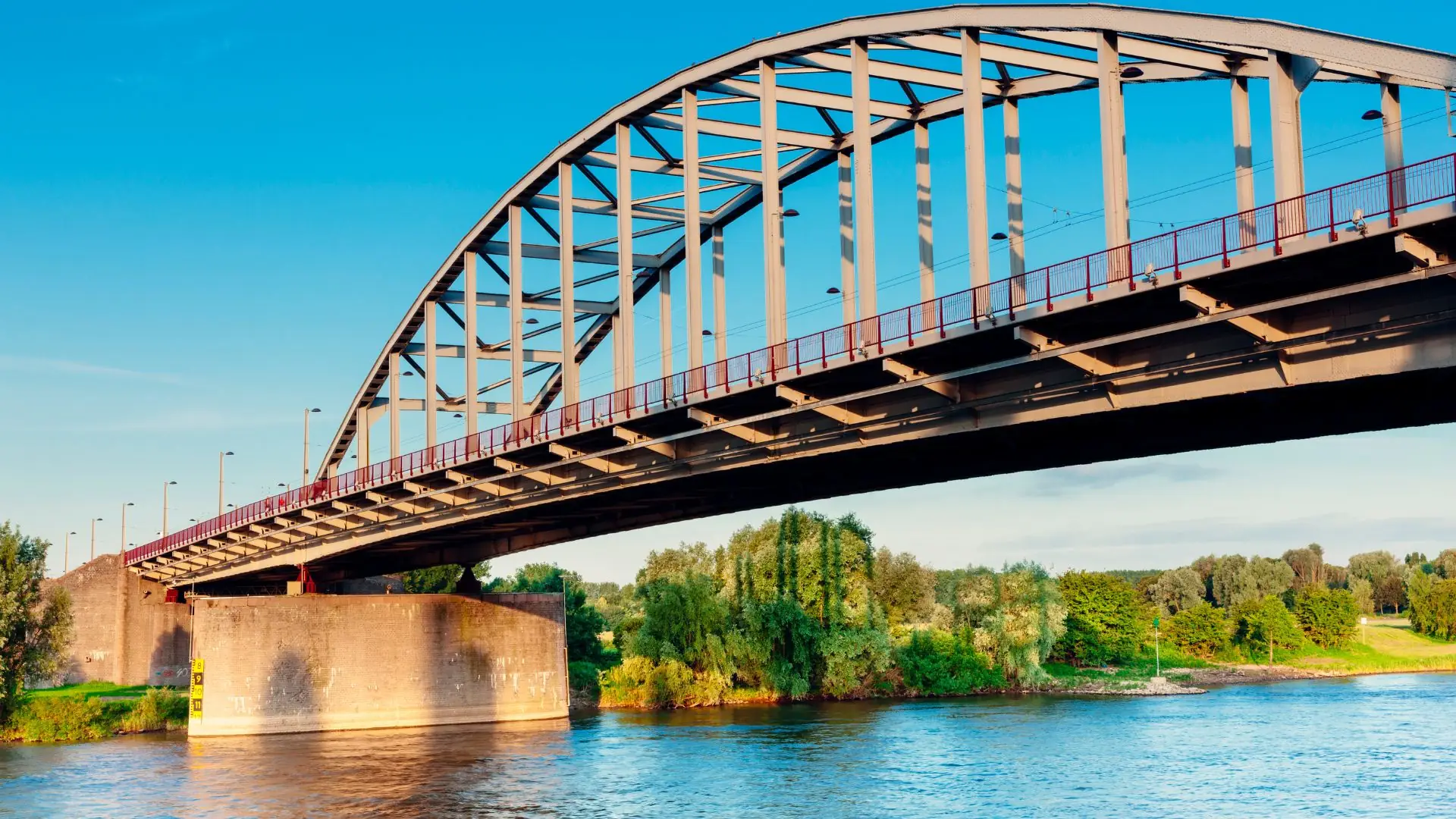 Stählerne Bogenbrücke über einen Fluss in Arnhem, umgeben von grüner Vegetation und blauem Himmel.