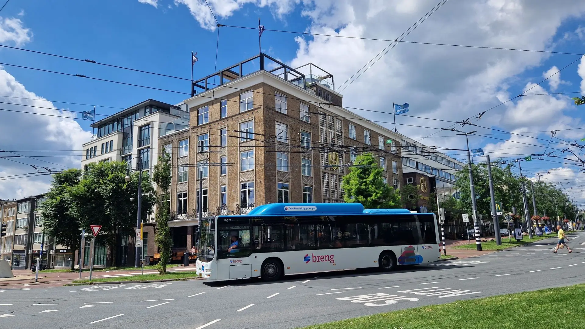 Blauer Bus der Marke Breng fährt an einer Kreuzung in Arnhem vorbei, im Hintergrund mehrstöckiges Gebäude und blauer Himmel mit Wolken.