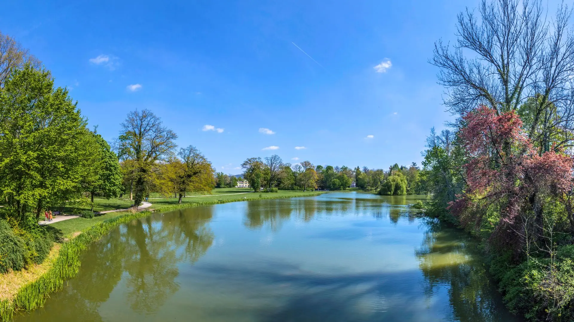 Blick auf einen breiten Fluss im Park Schönbusch in Aschaffenburg, umgeben von grünen und blühenden Bäumen unter blauem Himmel.
