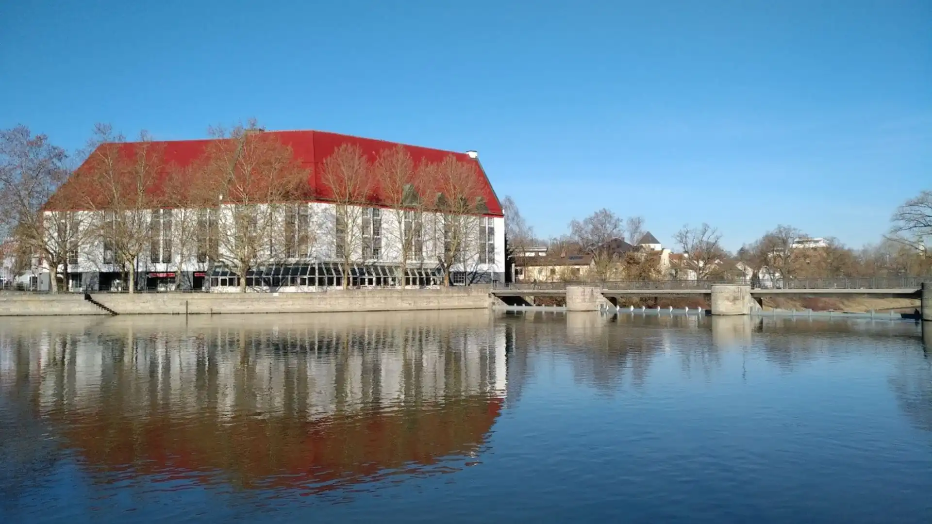 Außenansicht des Hotels Kaiserhof Landshut mit rotem Dach am Ufer eines ruhigen Flusses unter blauem Himmel