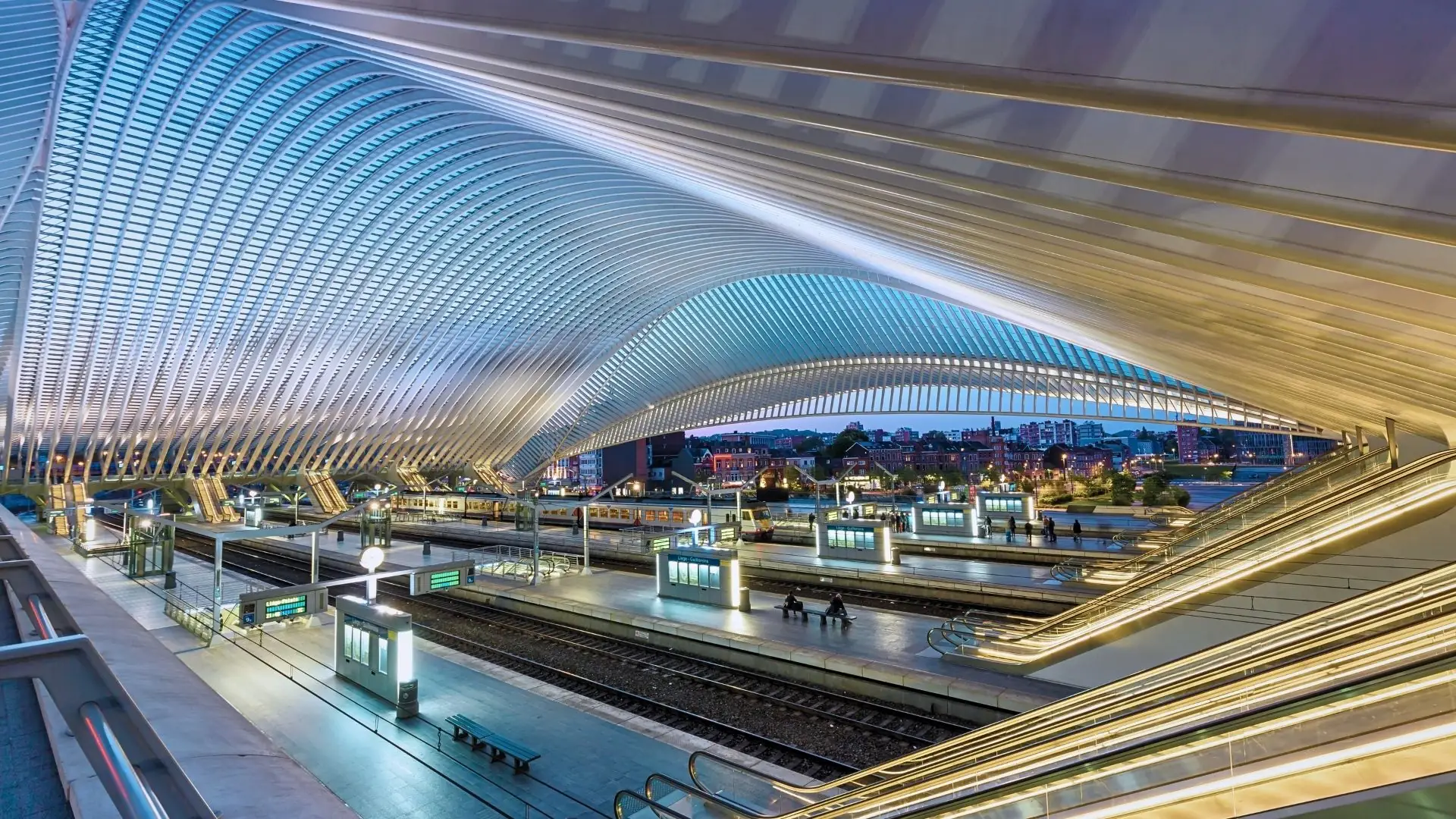 Blick auf den Bahnhof Liège-Guillemins mit geschwungener, lichtdurchlässiger Dachkonstruktion und Gleisen im Vordergrund, Abendstimmung.