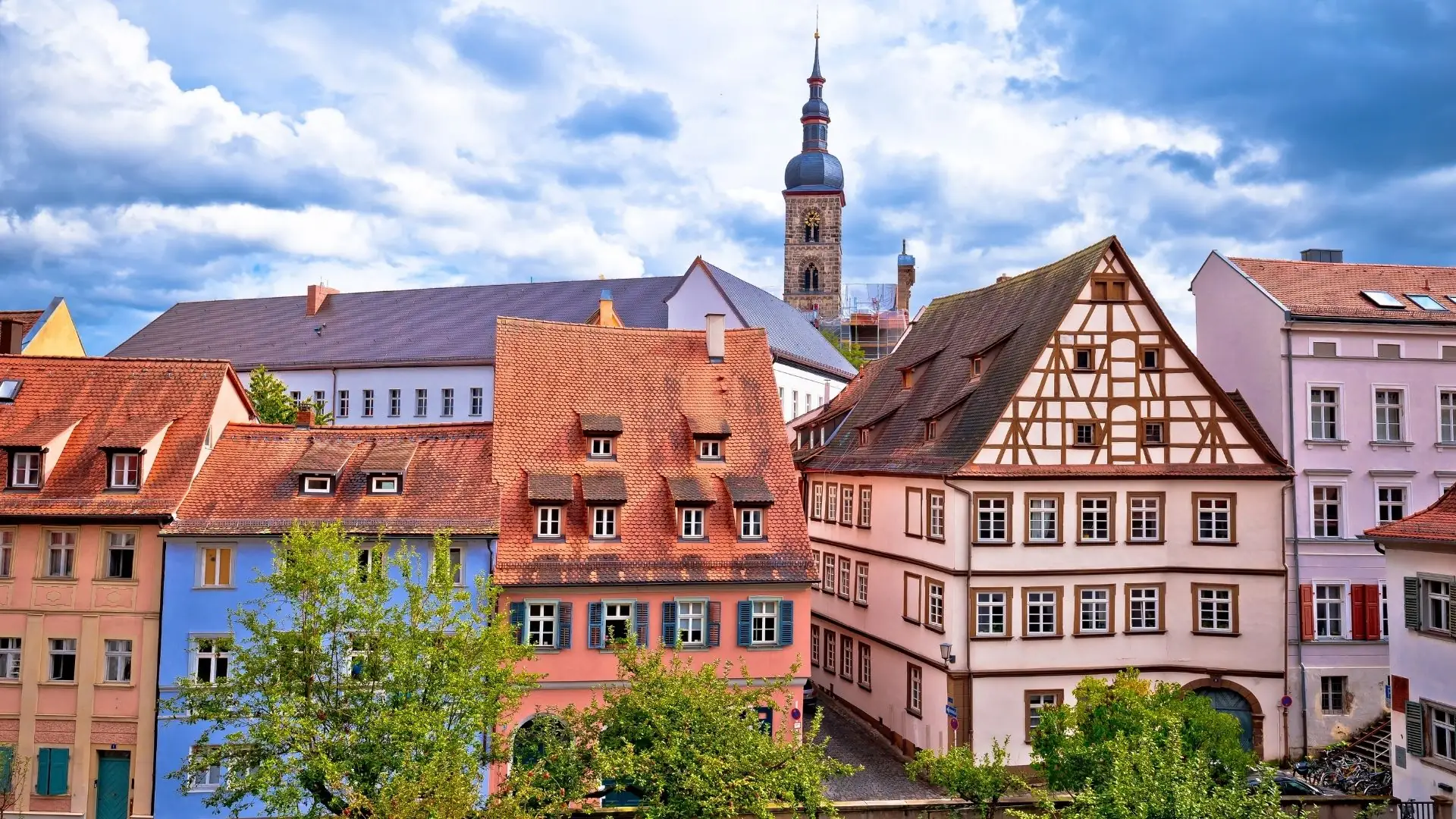 Bunte europäische Stadtansicht von Bamberg mit charmanten Fachwerkhäusern und roten Dächern. Im Hintergrund ragt ein Kirchturm unter einem teils bewölkten Himmel auf.