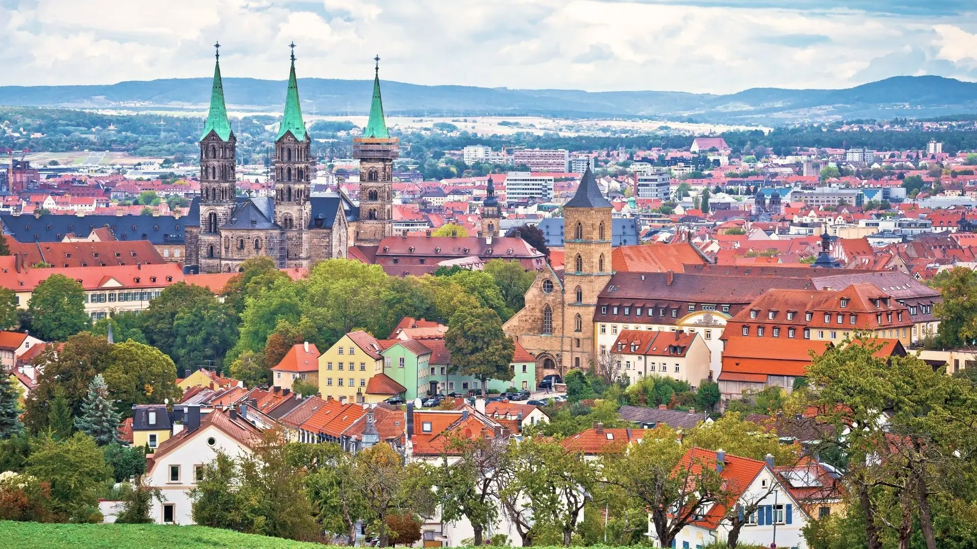 Ein Panoramablick auf Bamberg mit einer Skyline mit drei Türmen einer Kathedrale, umgeben von einer Mischung aus historischen Gebäuden mit orangefarbenen Dächern und üppigem Grün unter einem bewölkten Himmel.