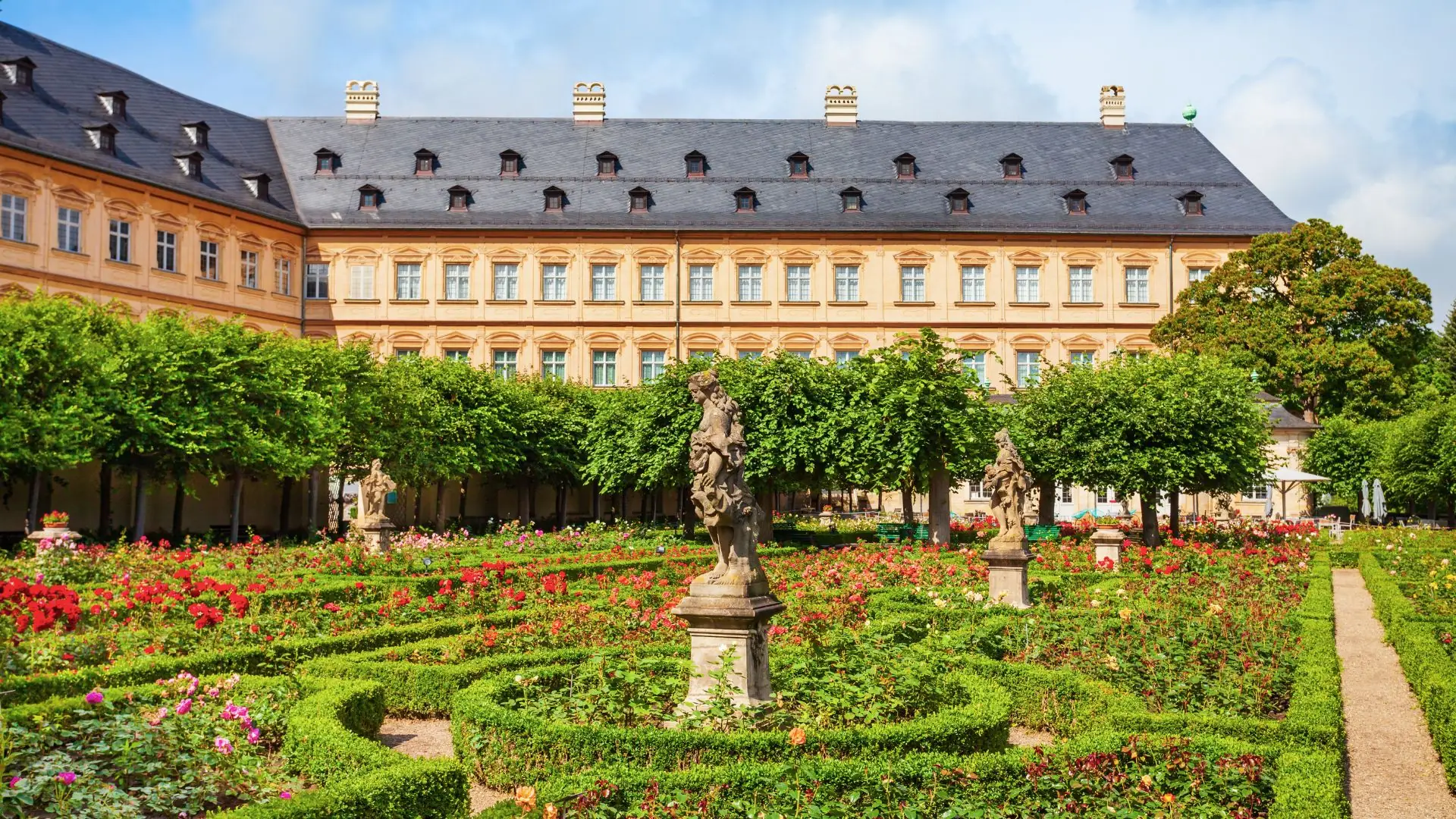 Barocke Neue Residenz in Bamberg mit symmetrischem Garten, Hecken, Blumenbeeten und mehreren steinernen Statuen auf Sockeln.