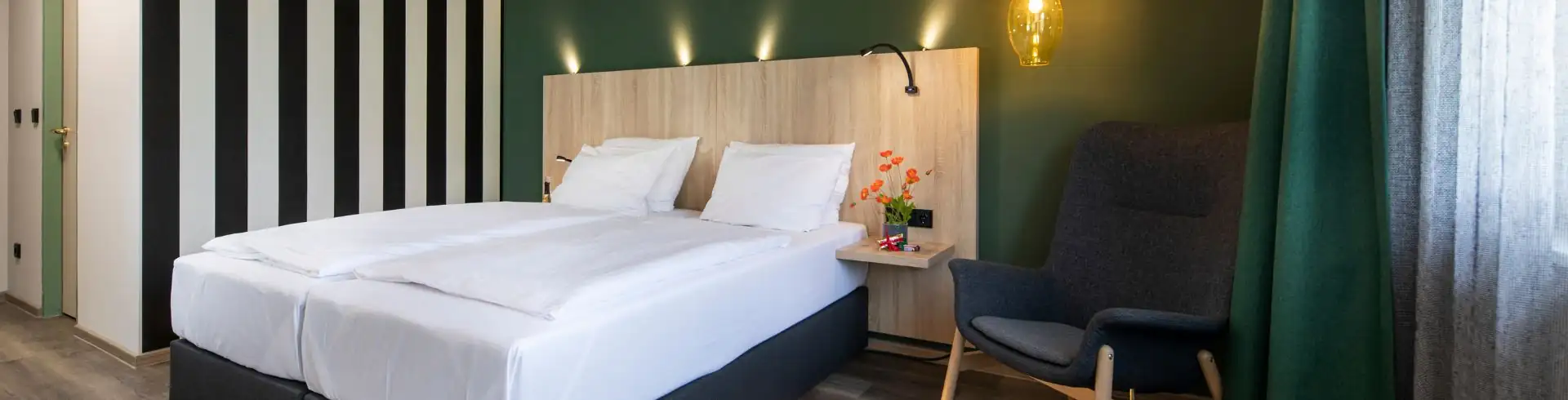 Modern eingerichtetes Hotelzimmer mit großem Doppelbett, Holz-Nachttisch, indirekter Beleuchtung unter dem Bett, grün gestrichener Wand. Banner