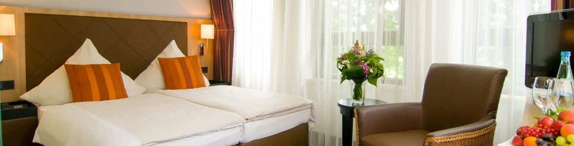 Banner Achat Hotel München Süd Gästezimmer mit Doppelbett, Sessel und Fernseher. Im Vordergrund steht ein frischer Obstkorb und eine Flasche Mineralwasser mit Gläsern. Durch die Vorhänge fällt warmes Tageslicht ins Zimmer. Neben dem Sessel steht ein Strauß frischer Schnittblumen.