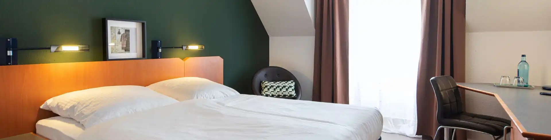 Banner. Hotelzimmer mit Doppelbett vor dunkelgrüner Wand, Nachttisch, Sessel neben Fenster mit Vorhängen und Schreibtisch mit Stuhl und Wasserflasche. Im ACHAT Hotel Heppenheim