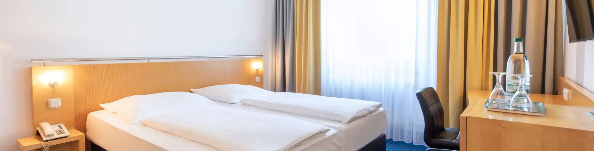 Komfortables Doppelzimmer im ACHAT Hotel Suhl mit großem Bett, Schreibtisch, Stuhl, Telefon und Fenster mit Vorhängen. Banner