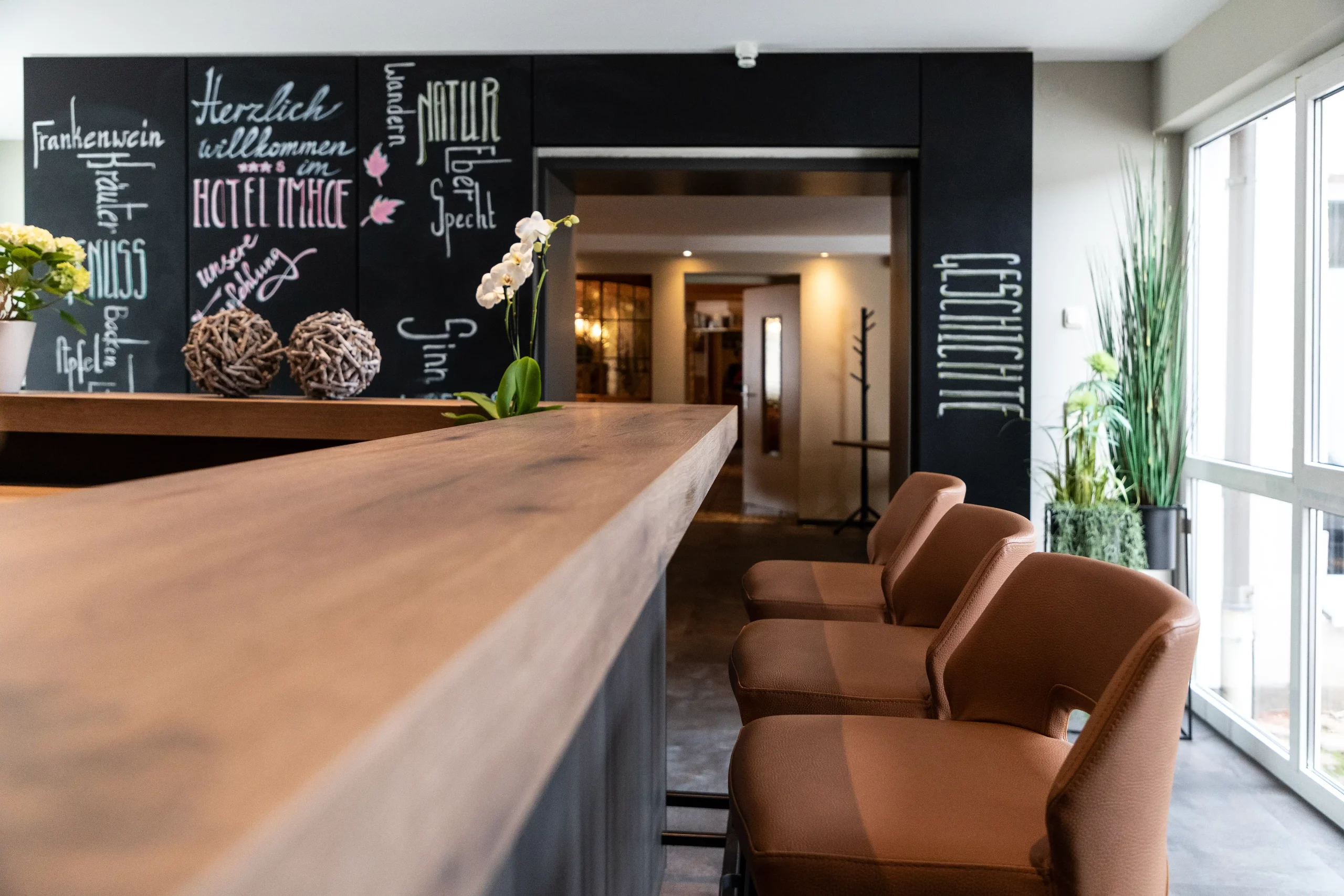 Barbereich im Hotel Imhof mit Holztheke, drei braunen Barhockern, schwarzer Wand mit weißer und rosa Schrift und großer Fensterfront mit Pflanzen