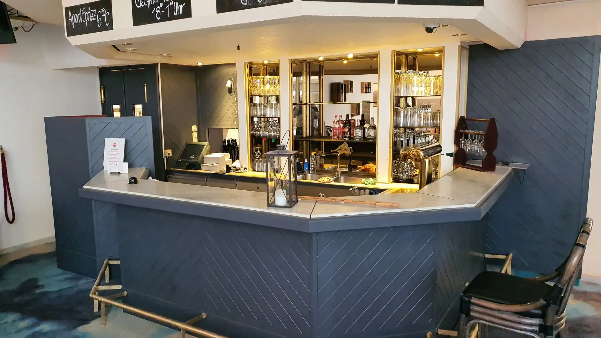 Bar mit hellgraßer Theke und dunklen Holzpaneelen, dahinter Regale mit Gläsern und Flaschen, davor zwei dunkle Stühle