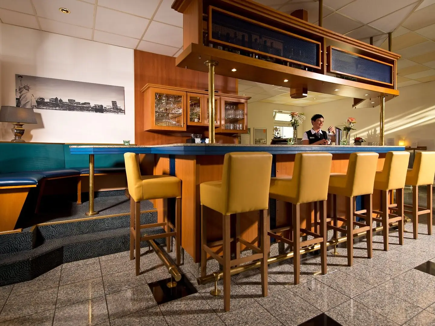 Barbereich mit Holztheke, gelben Barhockern und einer Person hinter der Theke im ACHAT Hotel Monheim am Rhein