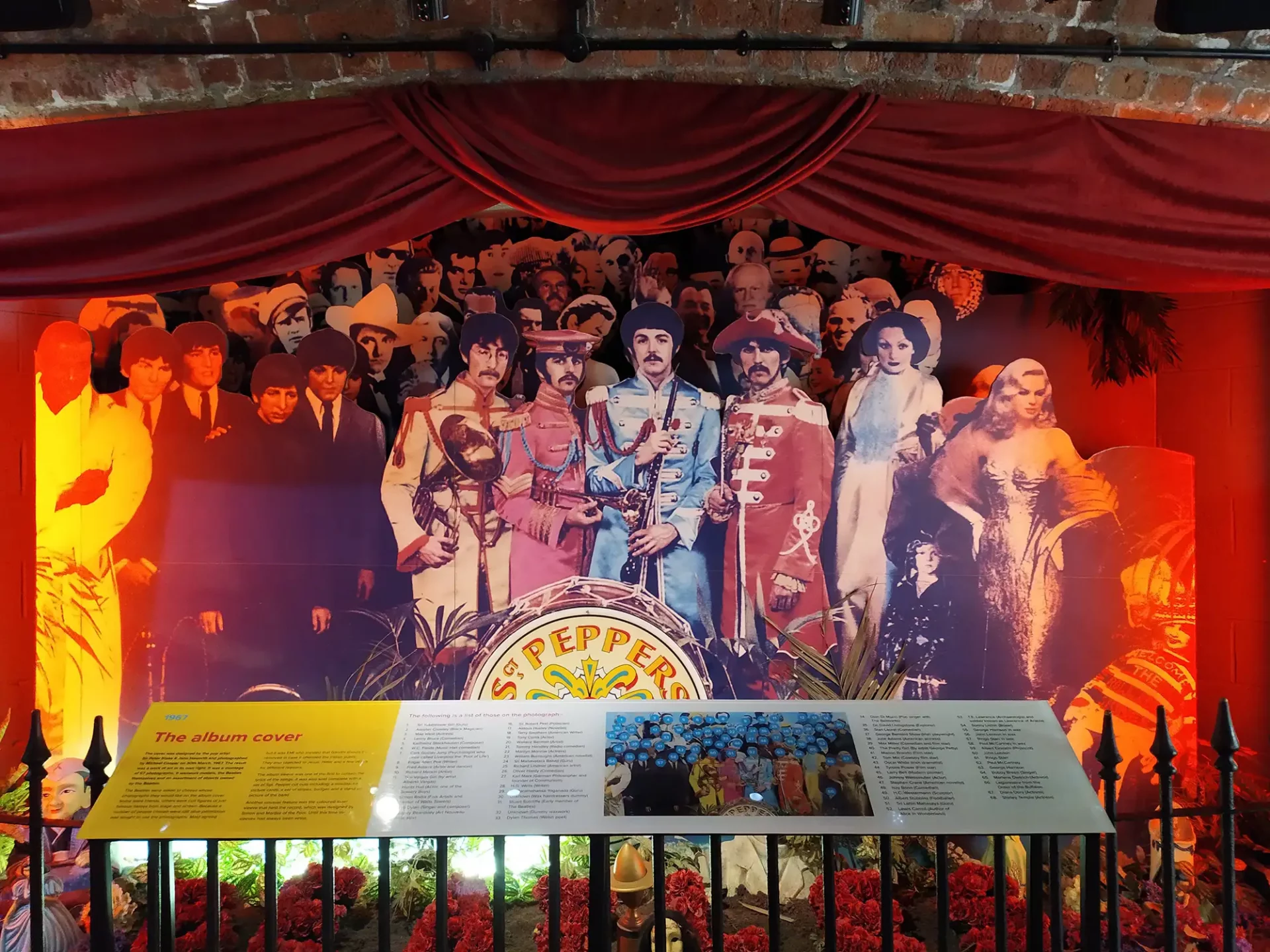 Beatles Museum Liverpool - Bild von den Beatles in bunten Kostümen vor einer Menschenmenge davor eine Infotafel und Blumen