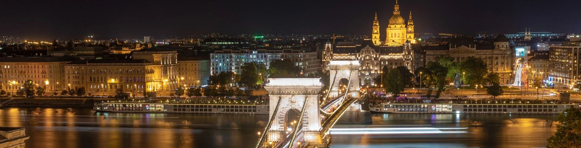 Städtereise Budapest zeigt die beleuchtete Kettenbrücke bei Nacht, die über die Donau führt, mit der eindrucksvoll erleuchteten St.-Stephans-Basilika und den historischen Gebäuden im Hintergrund.