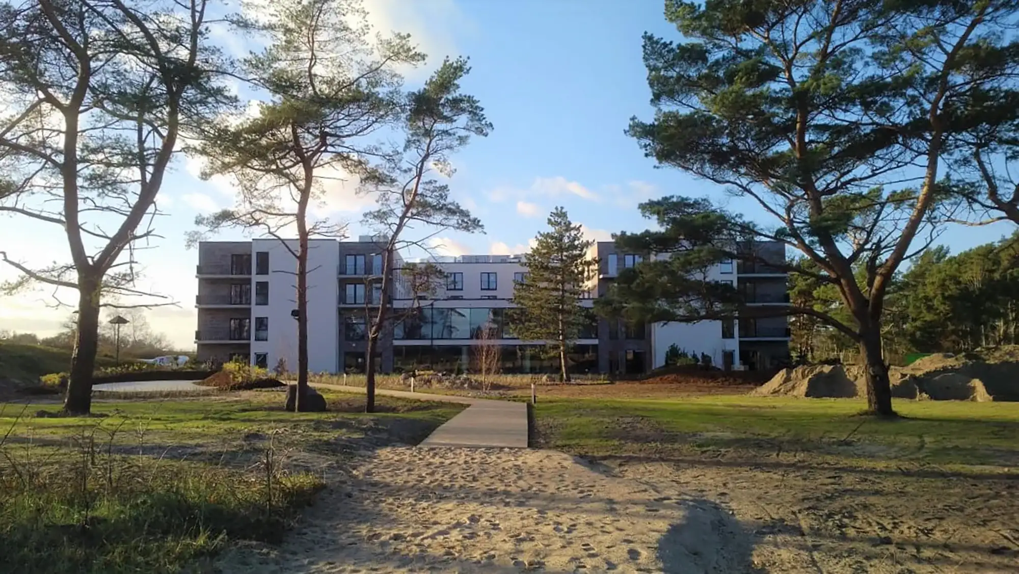 Blick vom Strand auf das Sante Royale Rügen Resort, umgeben von hohen Kiefern. Das moderne Gebäude fügt sich harmonisch in die naturnahe Umgebung ein, mit einem Holzsteg, der den Zugang über die Dünen ermöglicht.