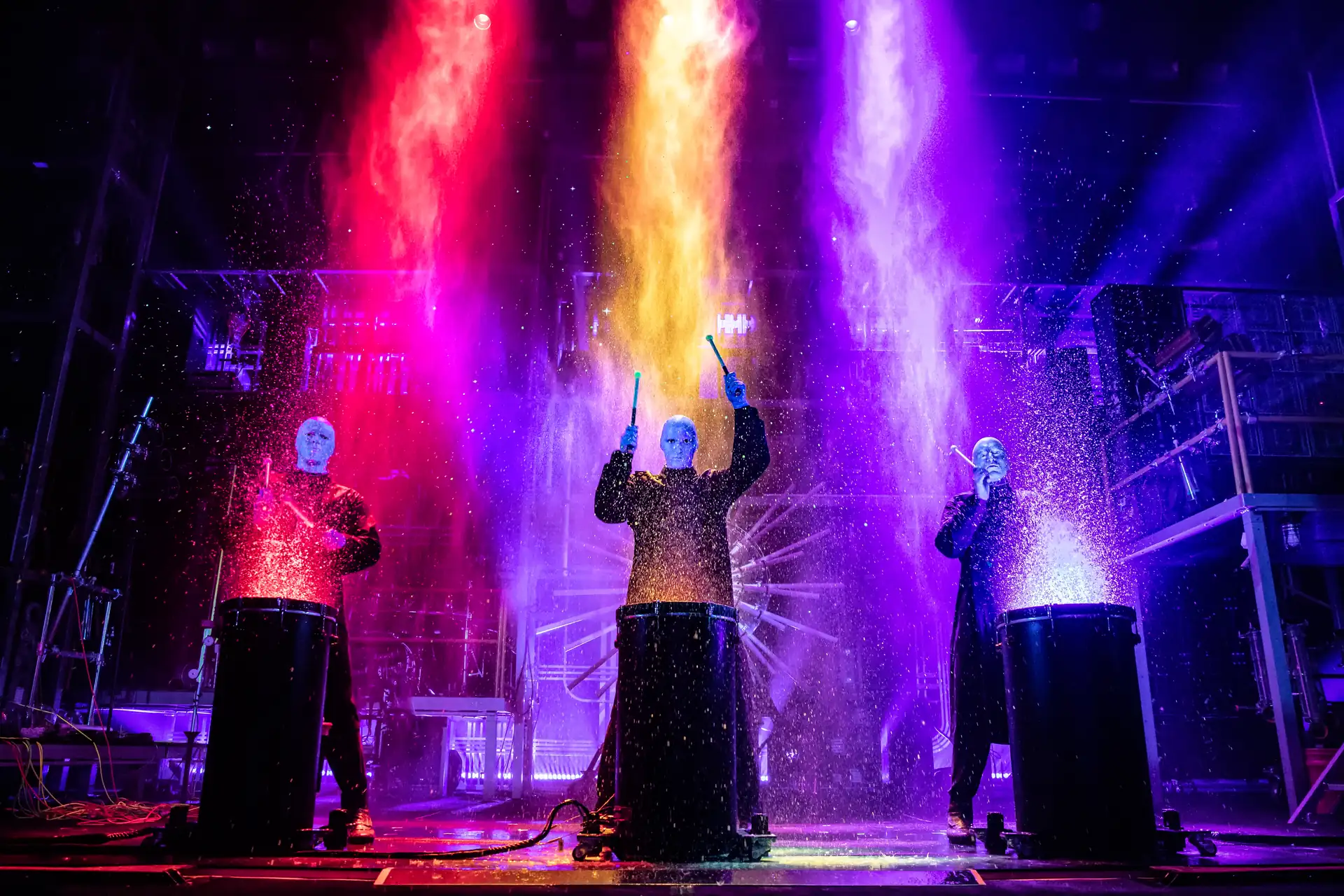 Drei Mitglieder der Blue Man Group stehen hinter großen Trommeln, aus denen farbige Pulver in Rot, Gelb und Blau aufsteigen, während sie mit Trommelstöcken spielen.