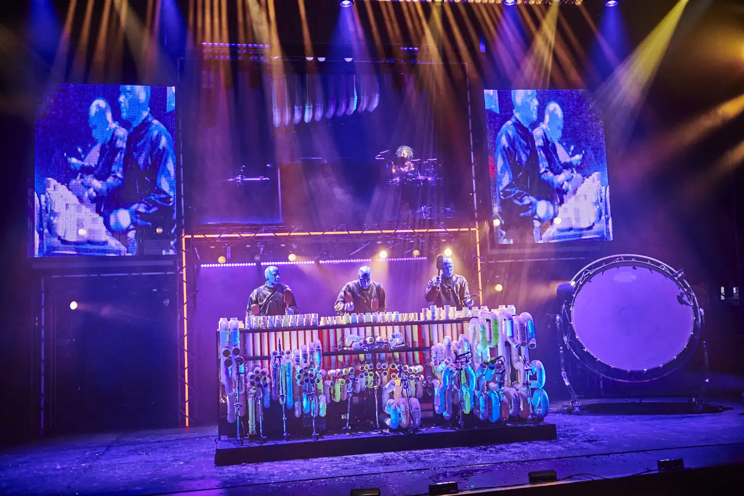 Drei Mitglieder der Blue Man Group mit blauer Gesichts- und Kopfbemalung spielen vor einer großen, bunten Perkussionsinstallation auf einer Bühne mit zwei großen Bildschirmen und einem großen Trommelfell im Hintergrund.