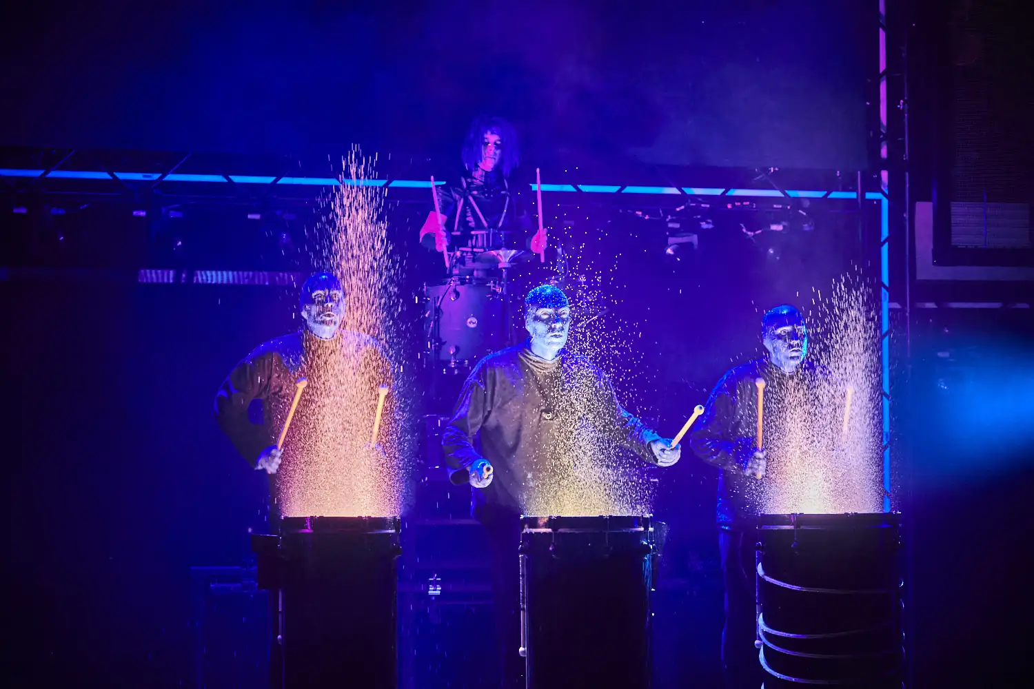 Drei Mitglieder der Blue Man Group schlagen auf Fässer, aus denen Funken sprühen, während ein Schlagzeuger im Hintergrund spielt.
