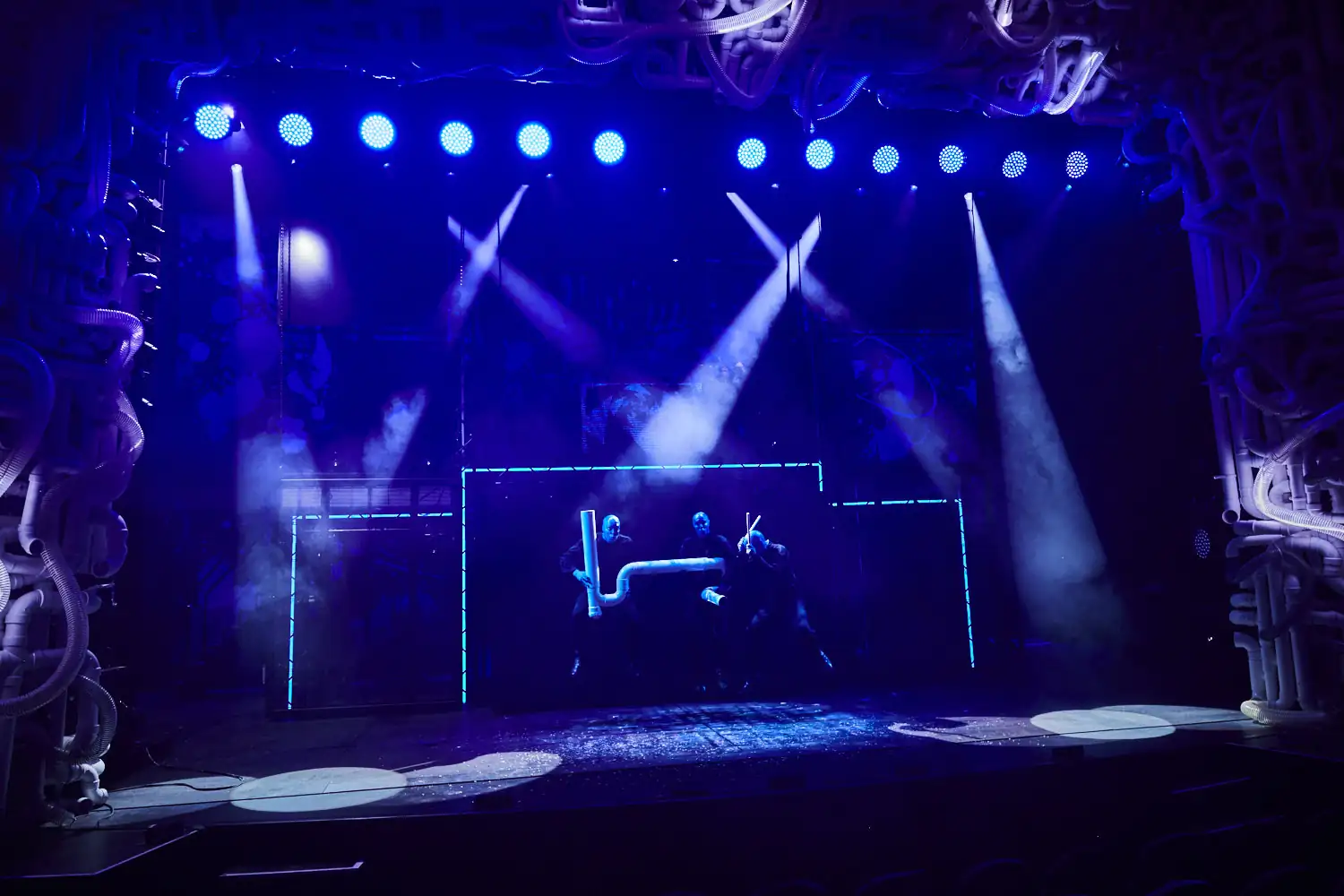 Bühne der Blue Man Group mit drei blau beleuchteten Figuren hinter leuchtenden rechteckigen Rahmen und Scheinwerfern im Hintergrund