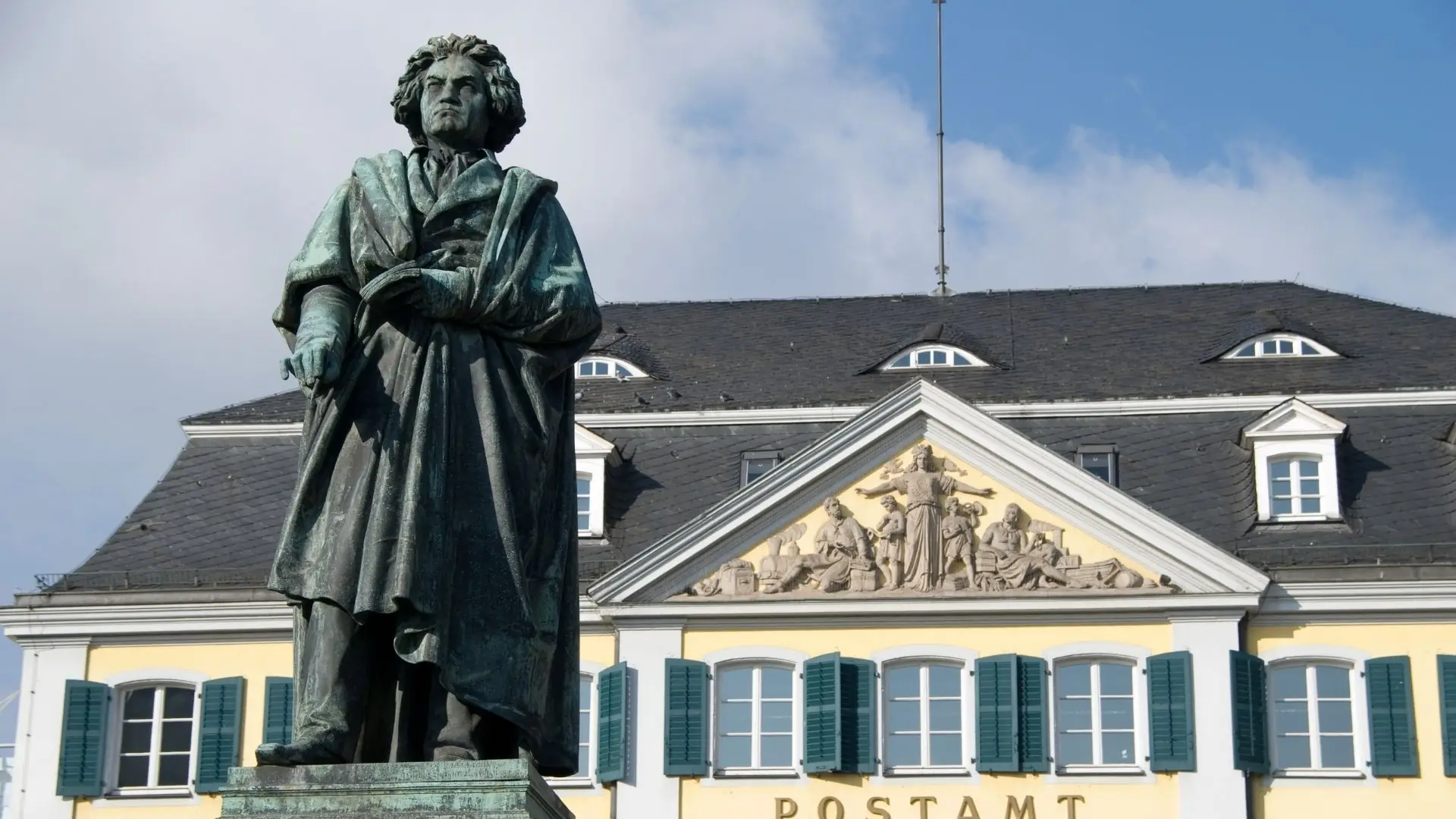 Bronzestatue von Ludwig van Beethoven, stehend mit dem Rücken zur Kamera, vor einem historischen Gebäude mit gelber Fassade und grünen Fensterläden.