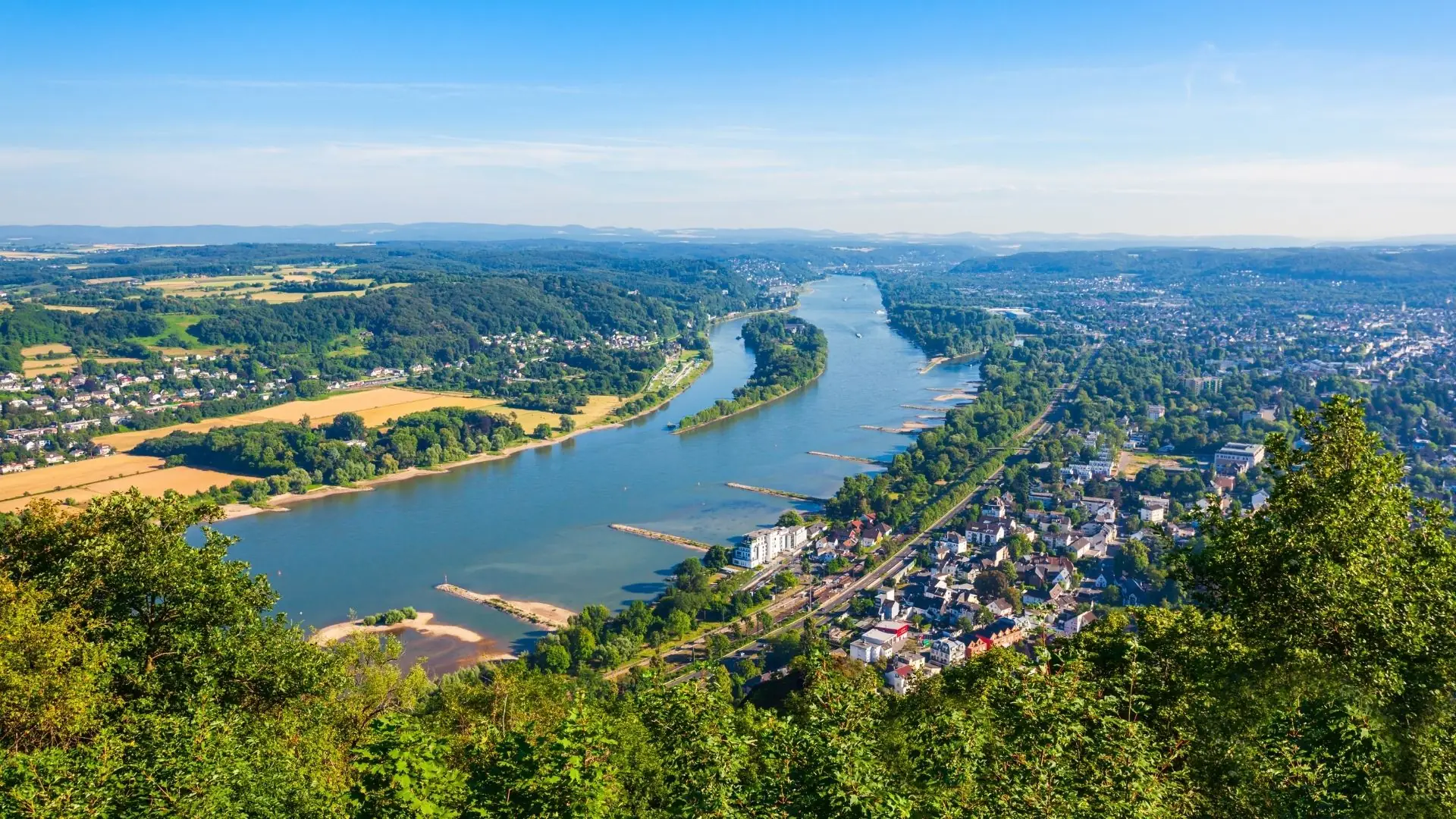 Panoramablick auf den Rhein bei Bonn, umgeben von grünen Hügeln und Feldern. Im Vordergrund sind Bäume und Häuser sichtbar.