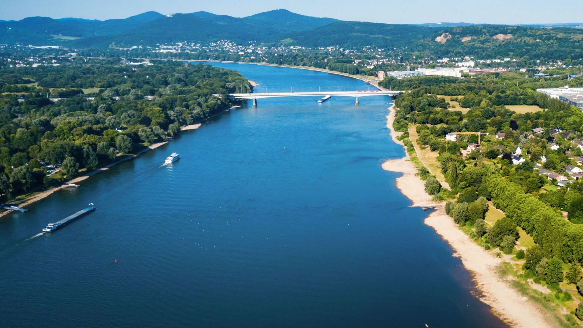 Luftaufnahme des Rheins in Bonn, mit einer Brücke, Booten auf dem Wasser und bewaldeten Uferbereichen.