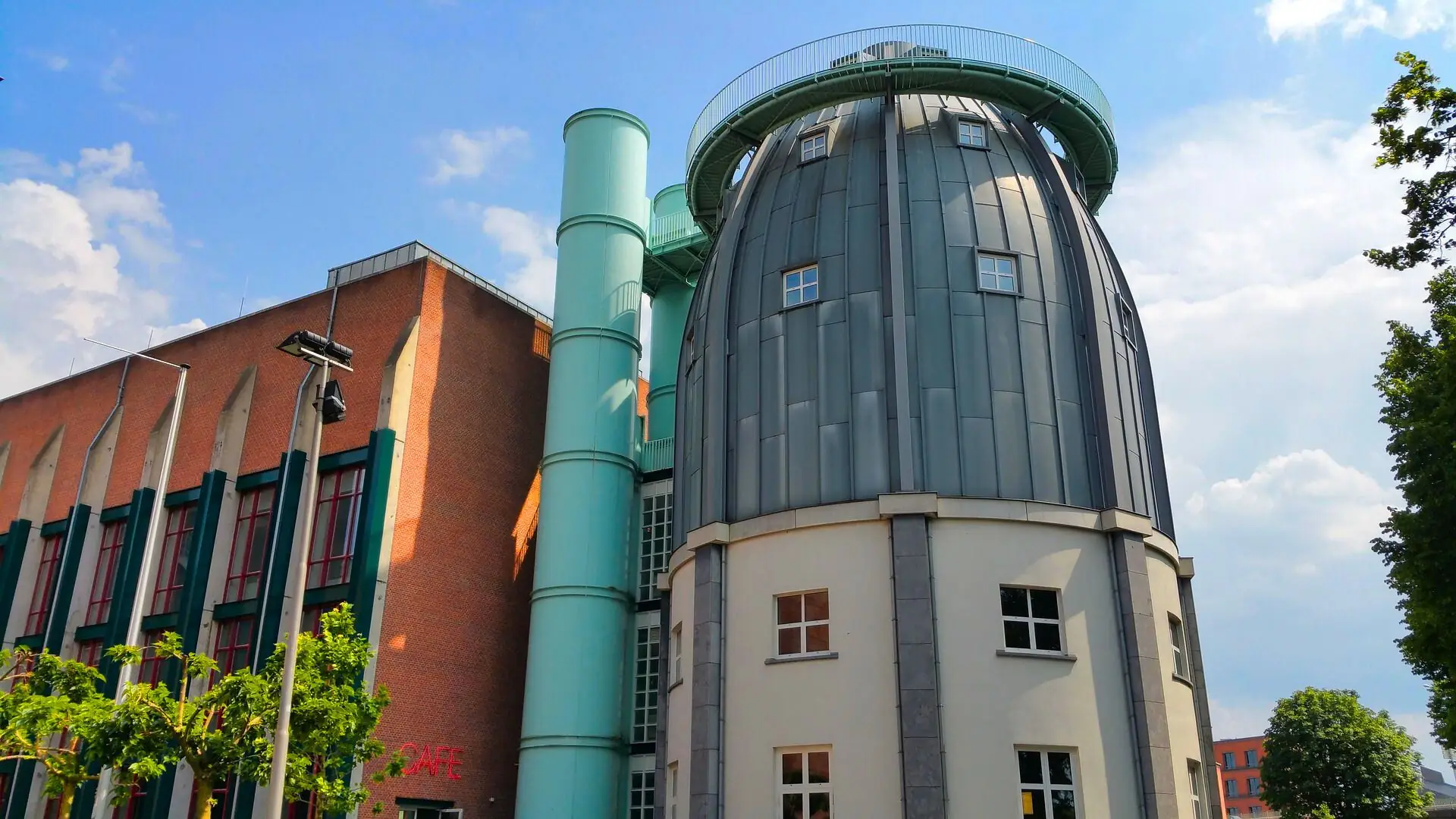 Rundes, graues Observatorium mit Fenstern neben rotem Backsteingebäude und grünem Lüftungsschacht unter blauem Himmel im Bonnefantenmuseum Maastricht.