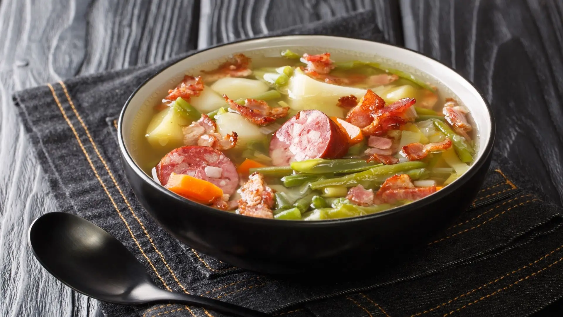 Luxemburger Bouneschlupp Gericht, in einer schwarzen Schale, darin grüne Bohnen, Kartoffelstücke, Karotten, Wurstscheiben und Speck, auf schwarzem Stoff mit orangefarbenen Nähten