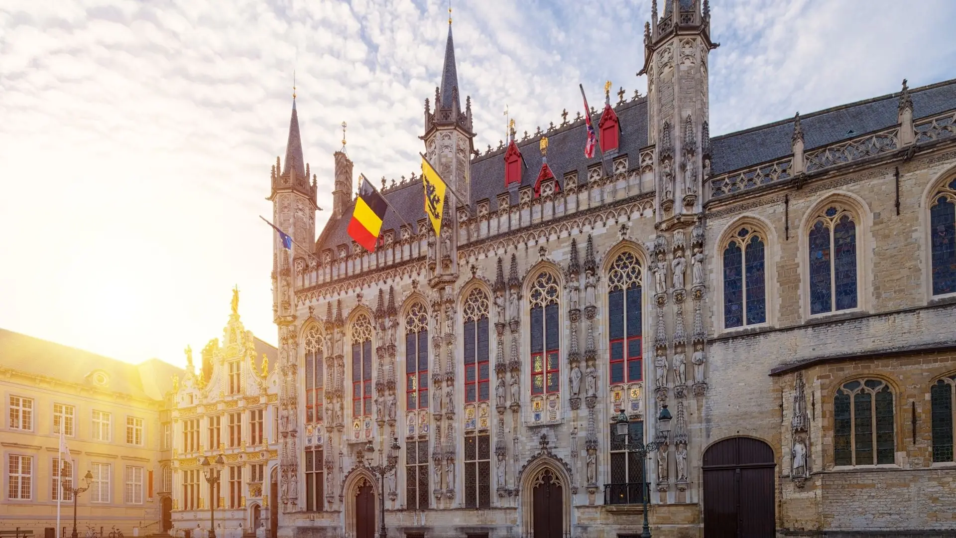 Das gotische Rathaus von Brügge mit hohen Türmen, verzierten Fenstern und Flaggen an der Fassade bei Sonnenuntergang