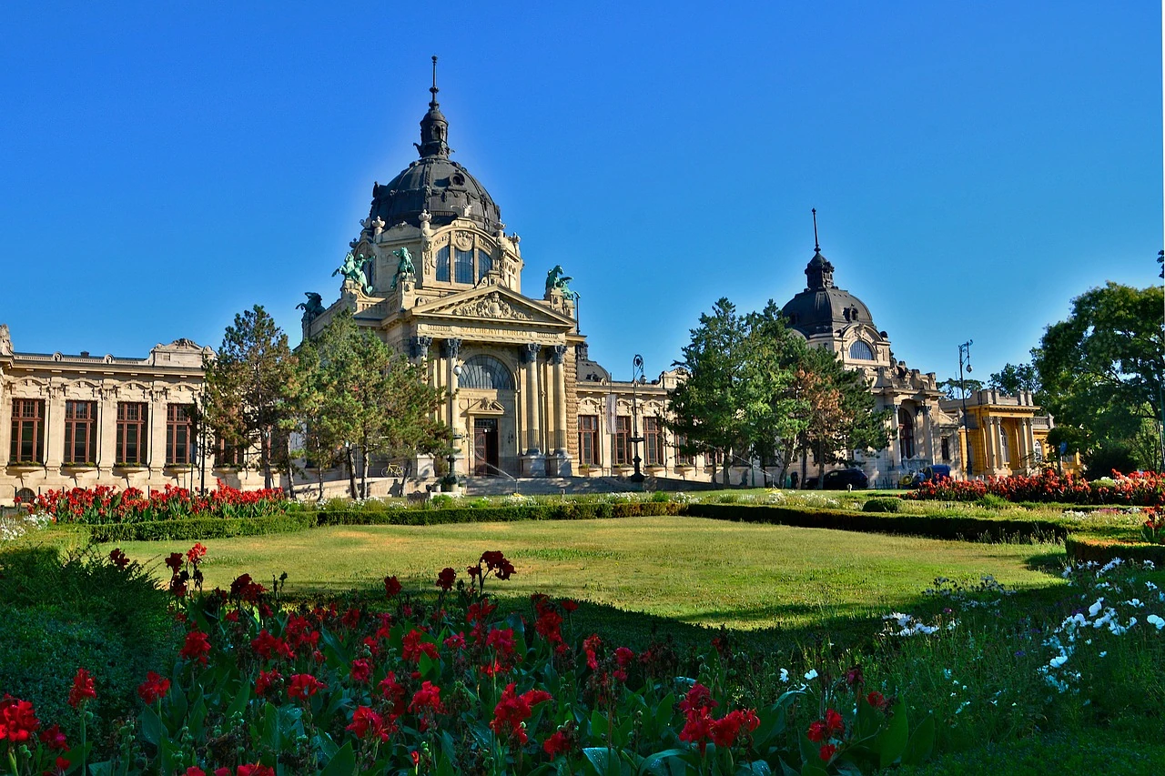 Szechenyi Thermalbad umgeben von gepflegten Gärten und blühenden Blumen, unter einem strahlend blauen Himmel.