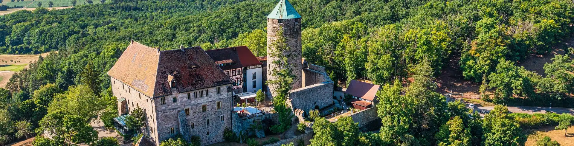 Burg Colmberg mit rundem Bergfried und umlaufender Mauer auf bewaldetem Hügel mit umliegender Landschaft Banner