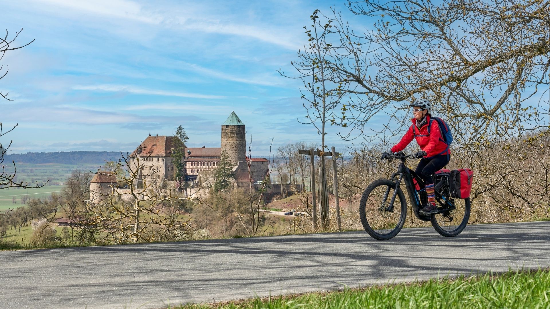 Radfahrerin mit roter Jacke und Helm fährt auf einem Weg vor der Burg Colmberg, die auf einem Hügel mit mehreren Türmen steht