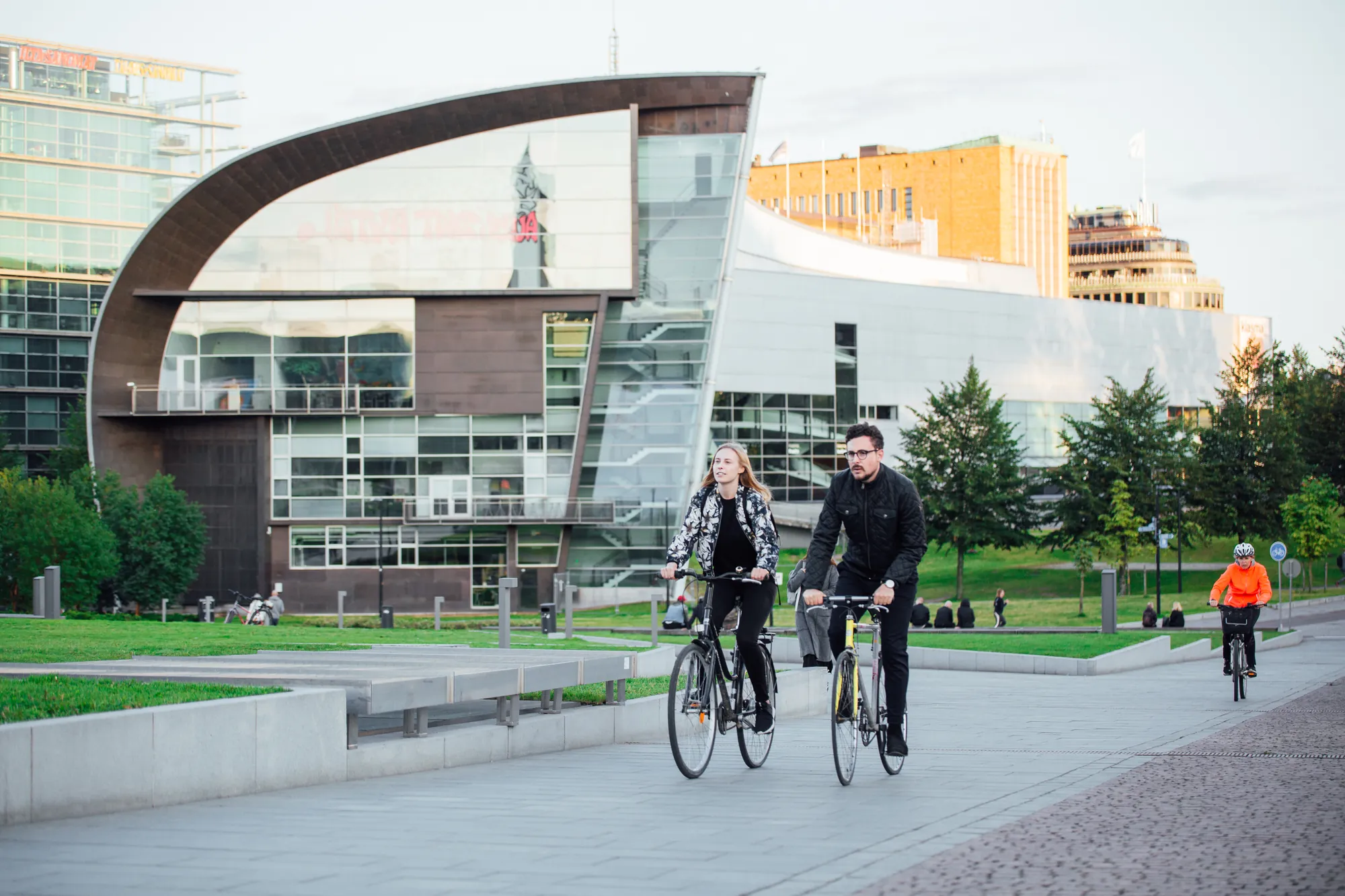 Zwei Fahrradfahrer vor dem Kiasma Museum in Helsinki, mit dem modernen, ästhetischen Museumsgebäude im Hintergrund bei Tageslicht, das durch seine geschwungene Architektur beeindruckt.