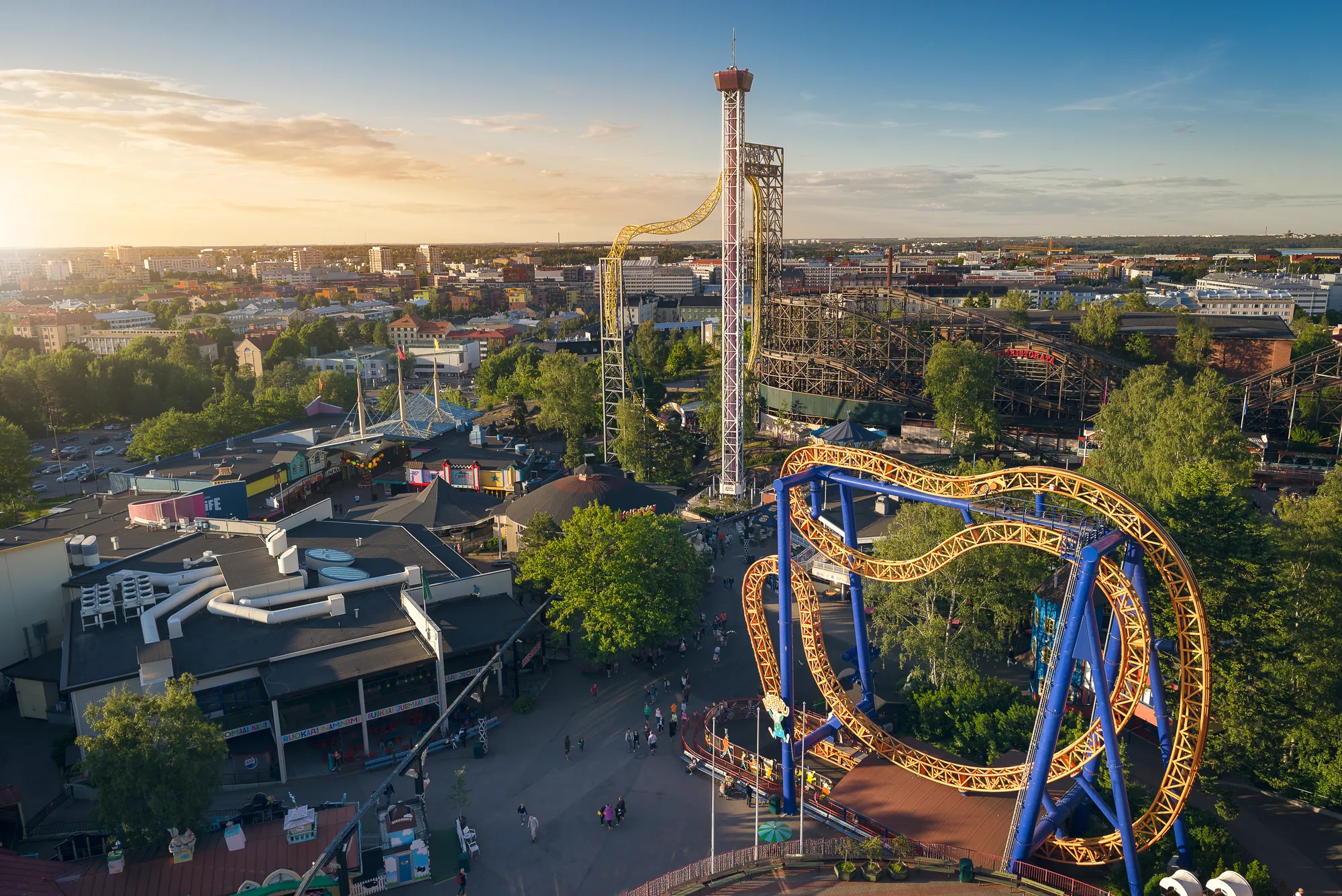 Luftaufnahme des Linnanmäki Freizeitparks, die verschiedene Attraktionen bei sonnigem Wetter zeigt, darunter Achterbahnen und Karussells, eingebettet in eine lebendige Parklandschaft.