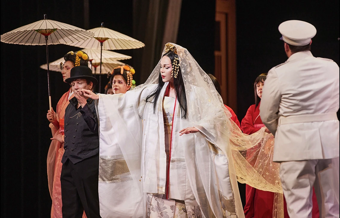 Theaterszene mit einer Frau in weißem, traditionellem japanischem Kimono und Schleier, umgeben von Darstellern in historischen Kostümen, darunter ein Mann in weißer Uniform und zwei Frauen mit Papier-Sonnenschirmen.