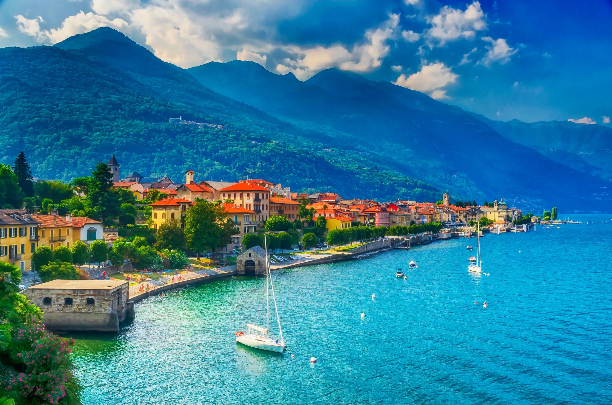 Cannobio am Lago Maggiore bei Tageslicht, mit dem glitzernden Wasser, auf dem einige Boote fahren. Bunte Häuser säumen die Promenade, wo Menschen entspannt in der Sonne sitzen. Im Hintergrund erheben sich majestätische Berge, die die malerische Kulisse vervollständigen.