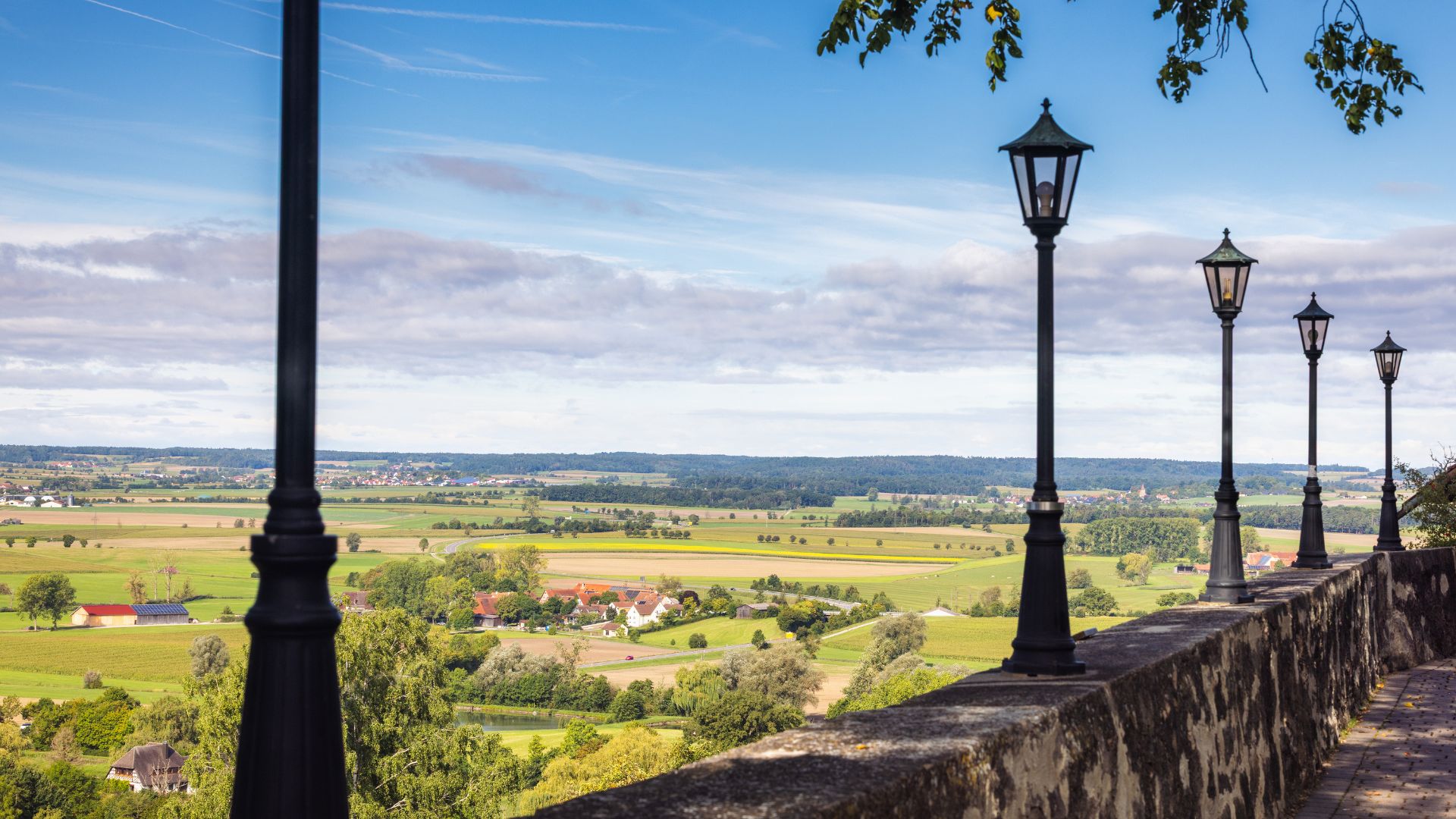 Blick von einer Steinmauer mit historischen Laternen auf eine weite, grüne Landschaft mit Feldern und vereinzelten Gebäuden bei Colmberg