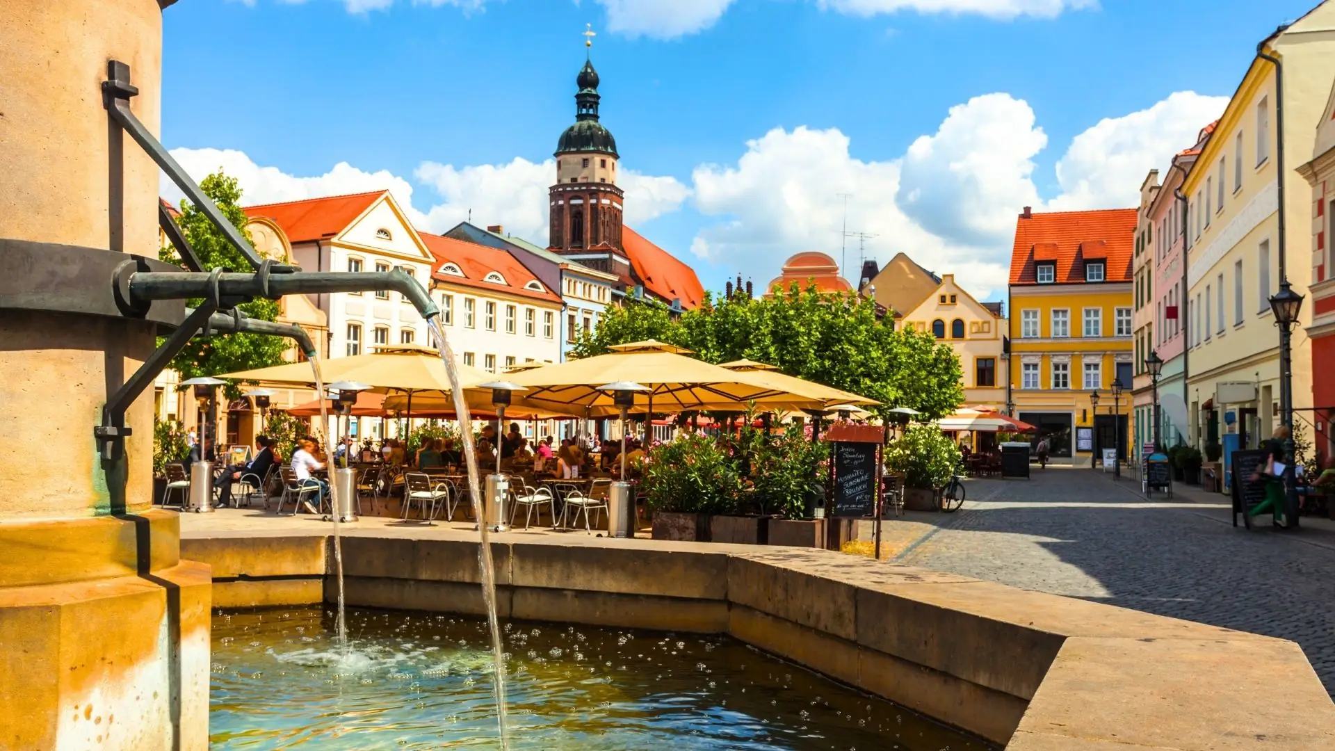Brunnen mit sprudelndem Wasser im Vordergrund, im Hintergrund historische Gebäude mit roten Dächern und einer Kirche mit Kuppel. Menschen sitzen unter Sonnenschirmen.