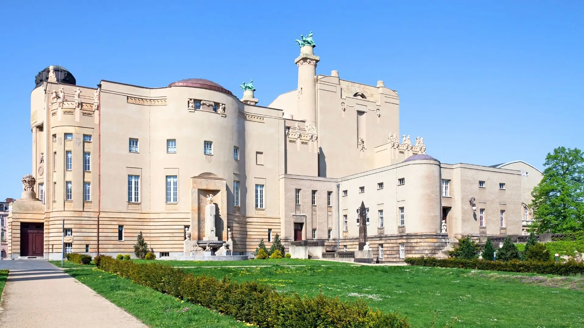 Das Staatstheater Cottbus, ein großes, historisches Gebäude mit einer markanten Fassade, zeigt eine Kombination aus klassizistischen und modernen Elementen. Im Vordergrund ist ein gepflegter Rasen mit Sträuchern zu sehen.