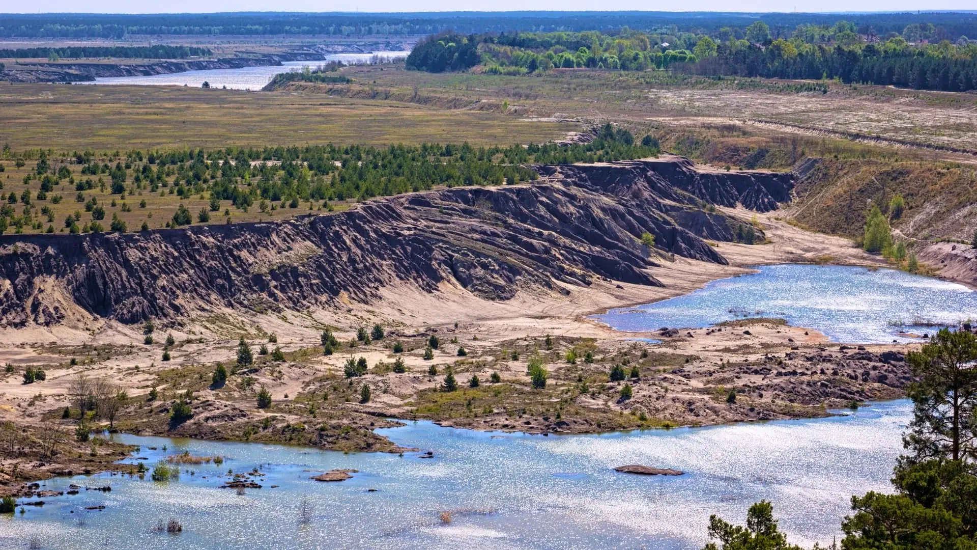 Landschaft mit sanften Hügeln, bewaldeten Bereichen und einem Fluss, der durch eine trockene, sandige Umgebung fließt. Im Vordergrund sind kleine Wasserflächen und bewachsene Hügel sichtbar.