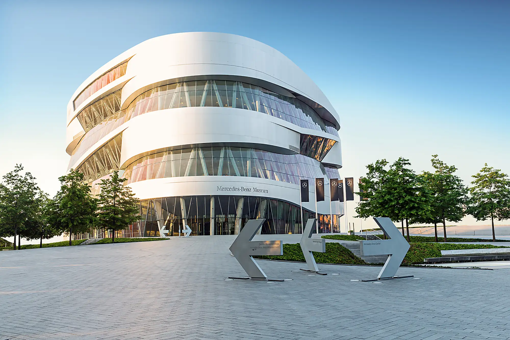 Außenaufnahme vom Mercedes Benz Museum mit Flaggen, Bäumen und Wegweisern als Pfeile