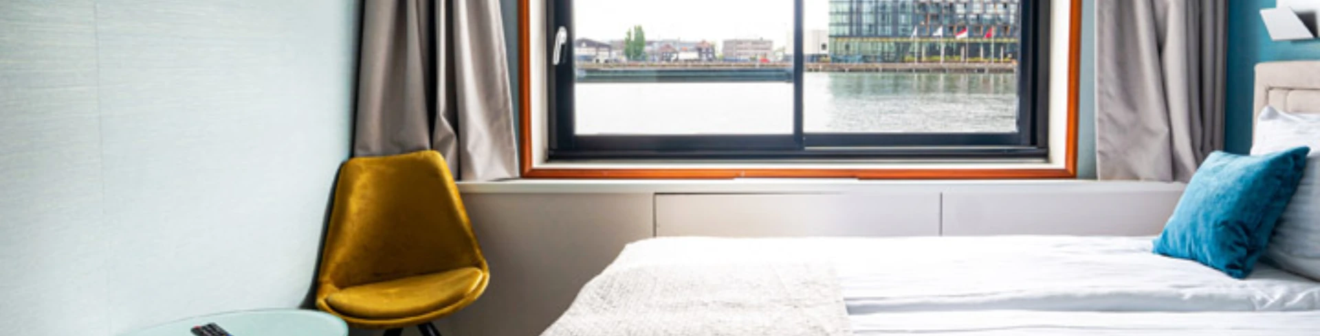 Hotelzimmer mit großem Fenster, Blick auf Wasser und Gebäude, Bett mit weißer Bettdecke und blauem Kissen, gelber Stuhl neben Tisch