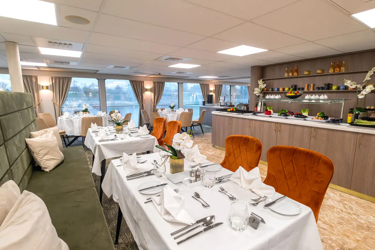Speisesaal mit gedeckten Tischen, weißen Tischdecken, orangen Stühlen und großen Fenstern mit Blick auf Wasser