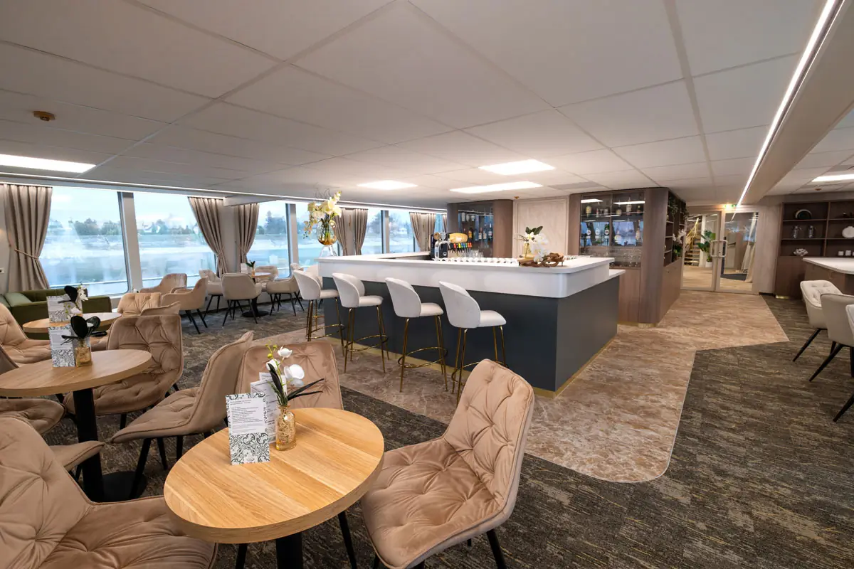 Moderne Hotelbar mit weißer Theke, fünf weißen Barhockern, runden Holztischen und gepolsterten Stühlen vor großen Fenstern mit Blick auf Wasser