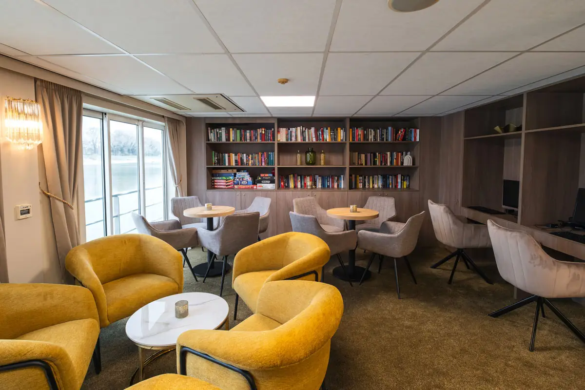 Gemütlicher Bibliotheksraum mit gelben Sesseln, runden Tischen, Bücherregalen und großen Fenstern mit Blick auf Wasser