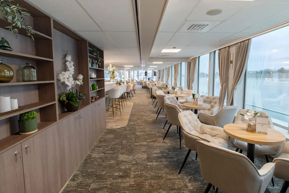 Moderner Speisesaal mit runden Holztischen, bequemen grauen Stühlen und großen Fenstern mit Blick auf Wasser